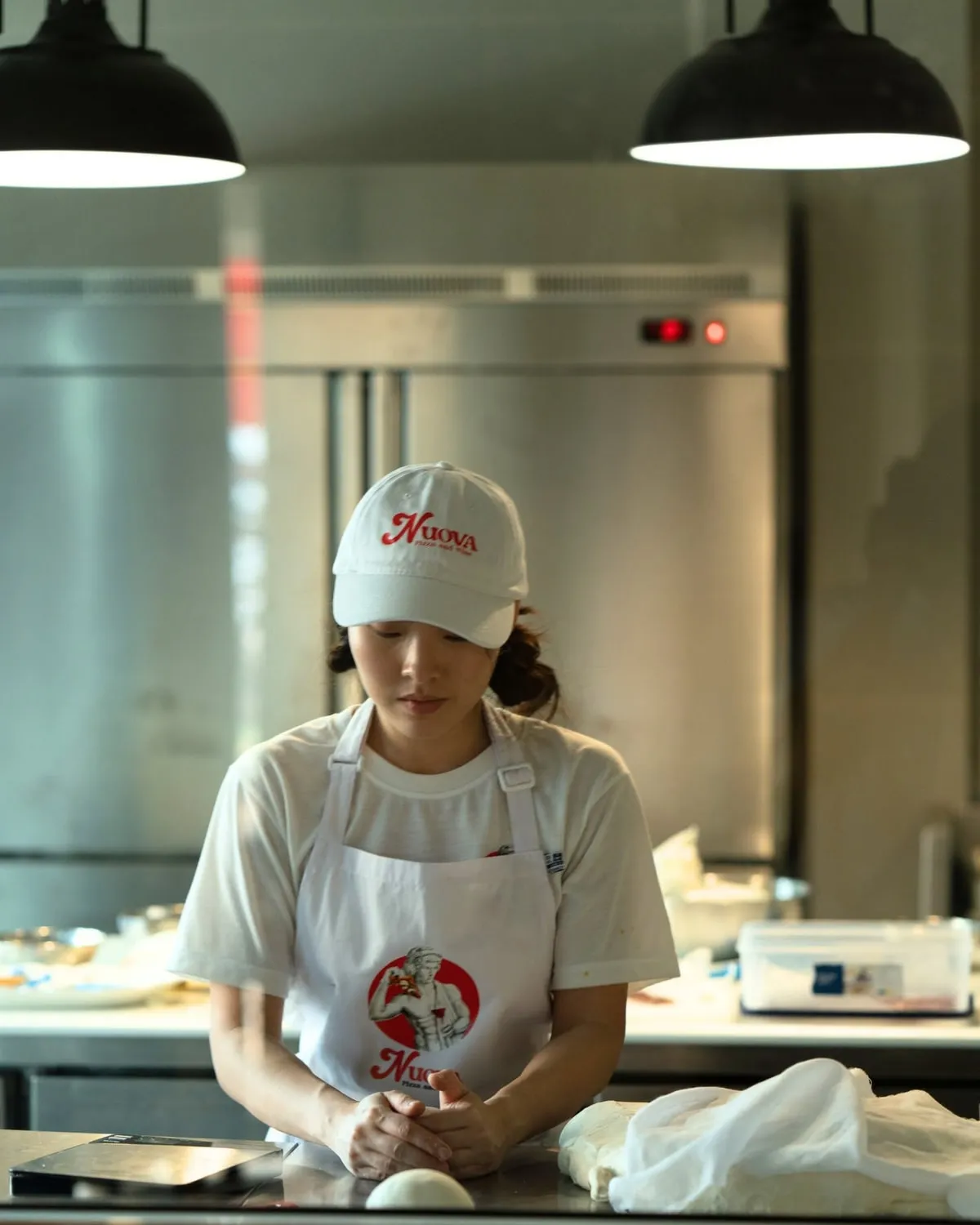 Nova at the steel bench in the Nuova kitchen, in a branded cap and apron, hands resting on a dough ball