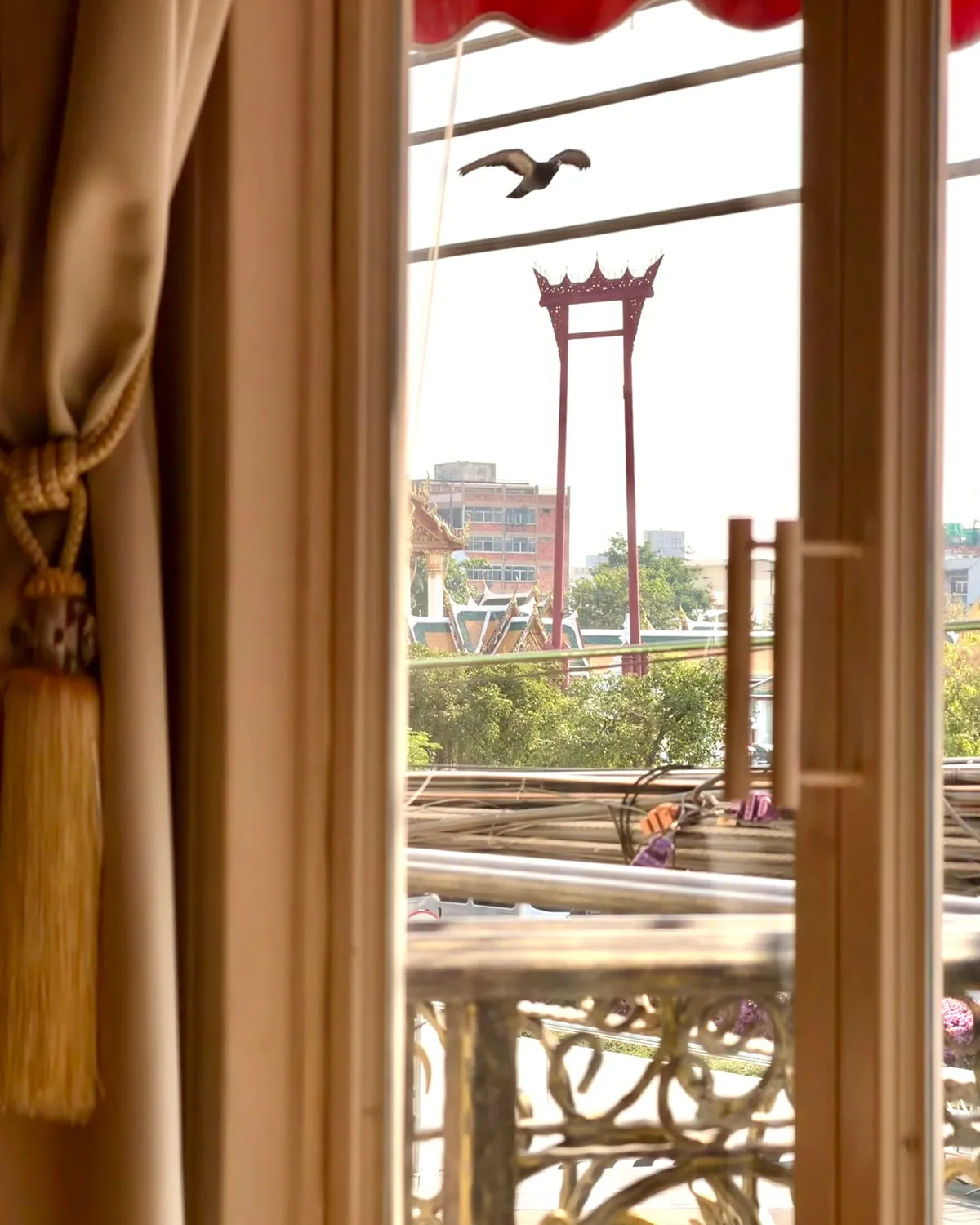 The Giant Swing seen from inside the restaurant, framed by a tied-back curtain and the balcony rail, a bird crossing above it