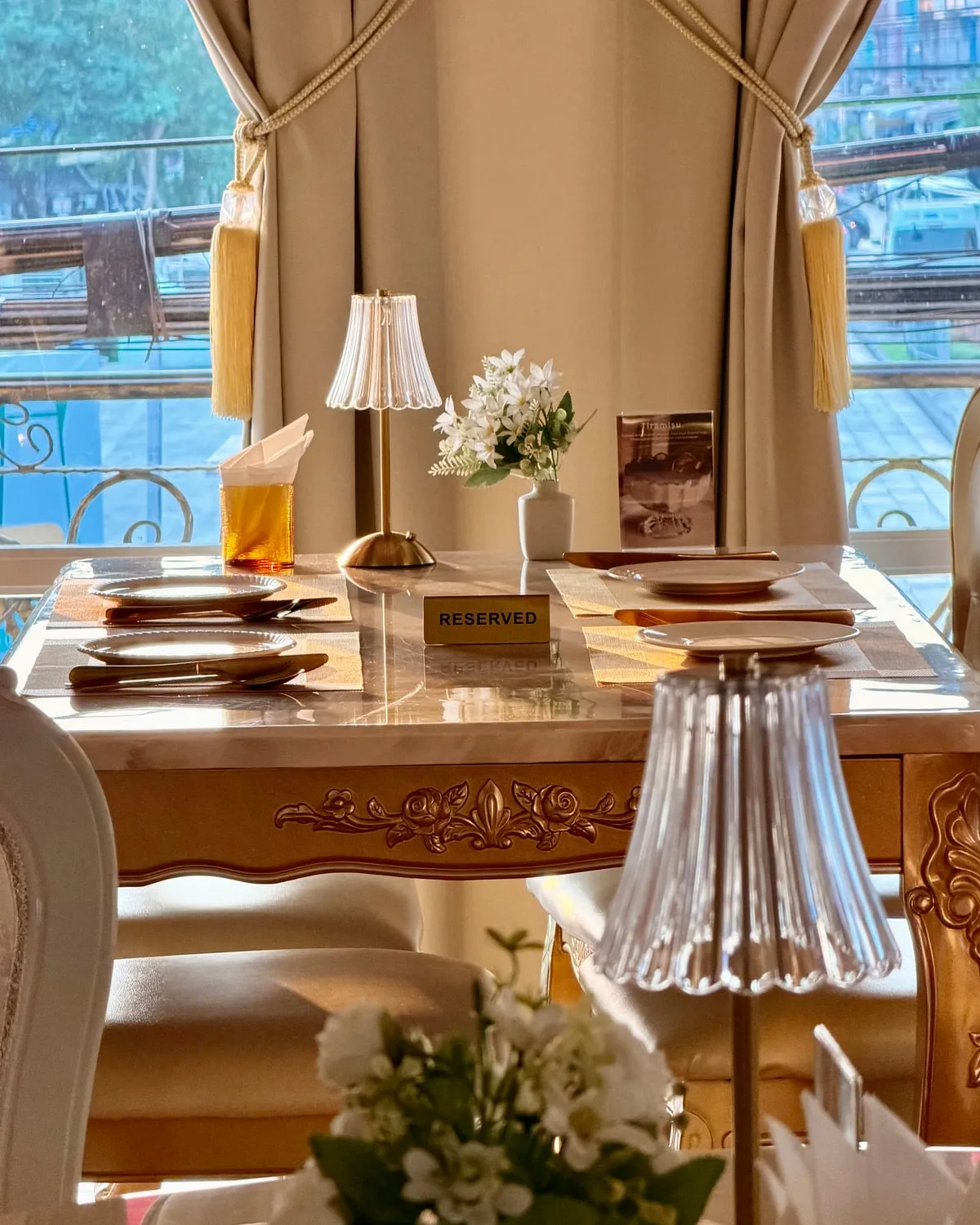 A table laid for two by the window in afternoon light, marble top on a carved gilt base, a small lamp and white flowers, a reserved card in the middle