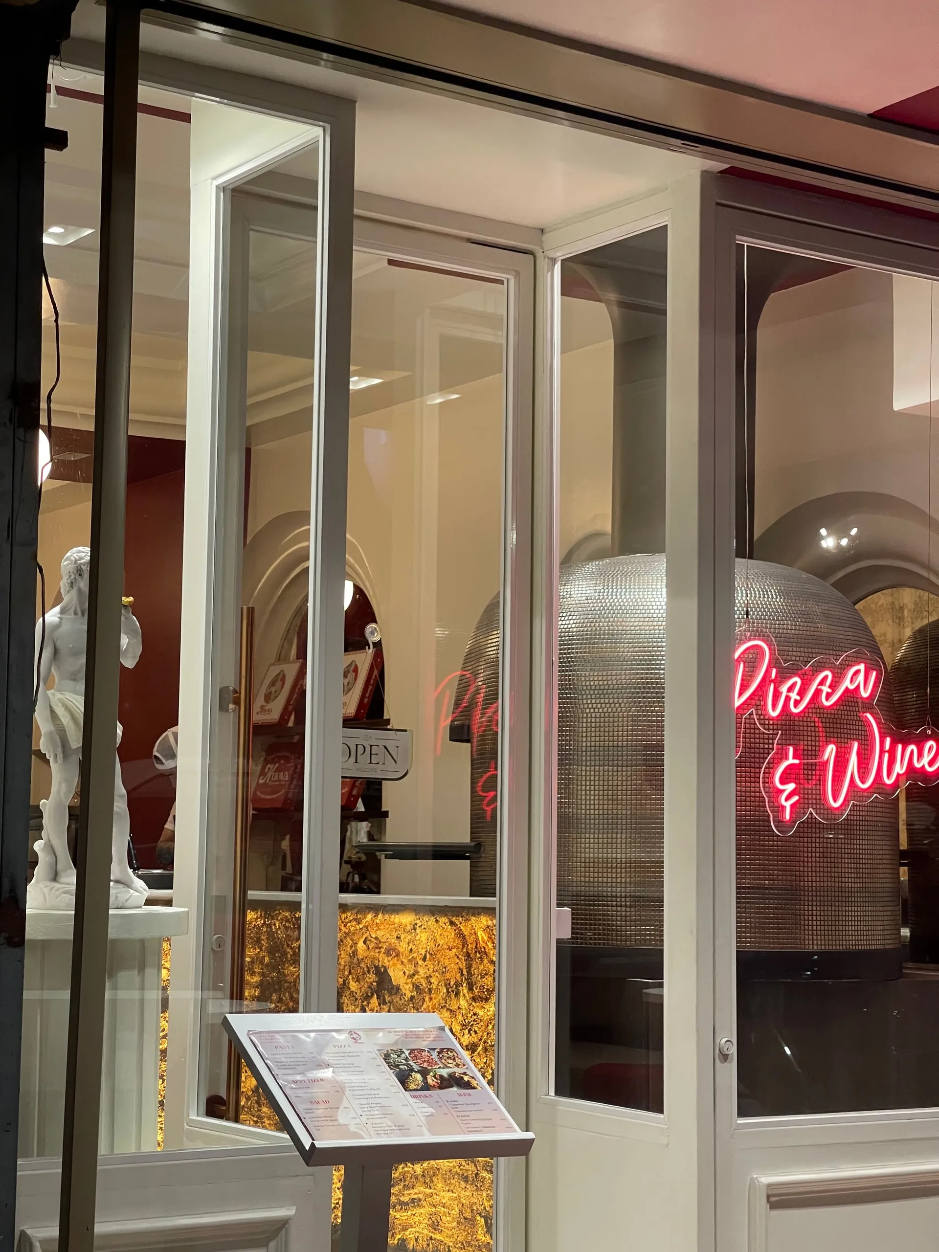 The Nuova shopfront after dark, the neon Pizza and Wine script glowing beside the mosaic oven and the David replica inside