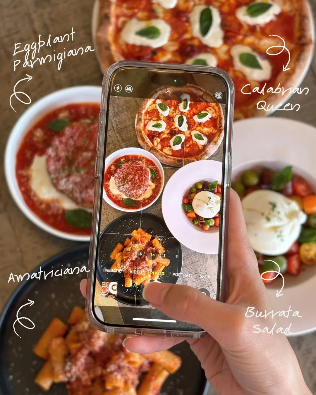 A table of dishes seen from above with a phone photographing them, hand-labelled Eggplant Parmigiana, Calabrian Queen, Amatriciana and Burrata Salad